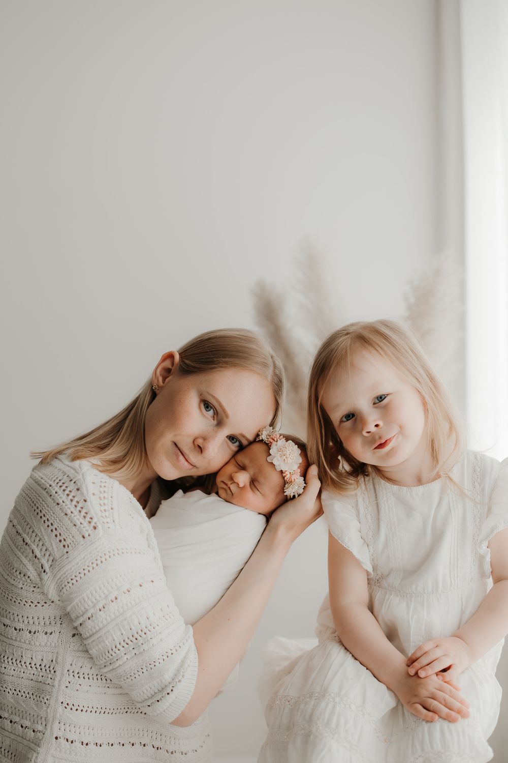 Mother in white knit top poses with newborn baby and young blonde daughter in bright studio.