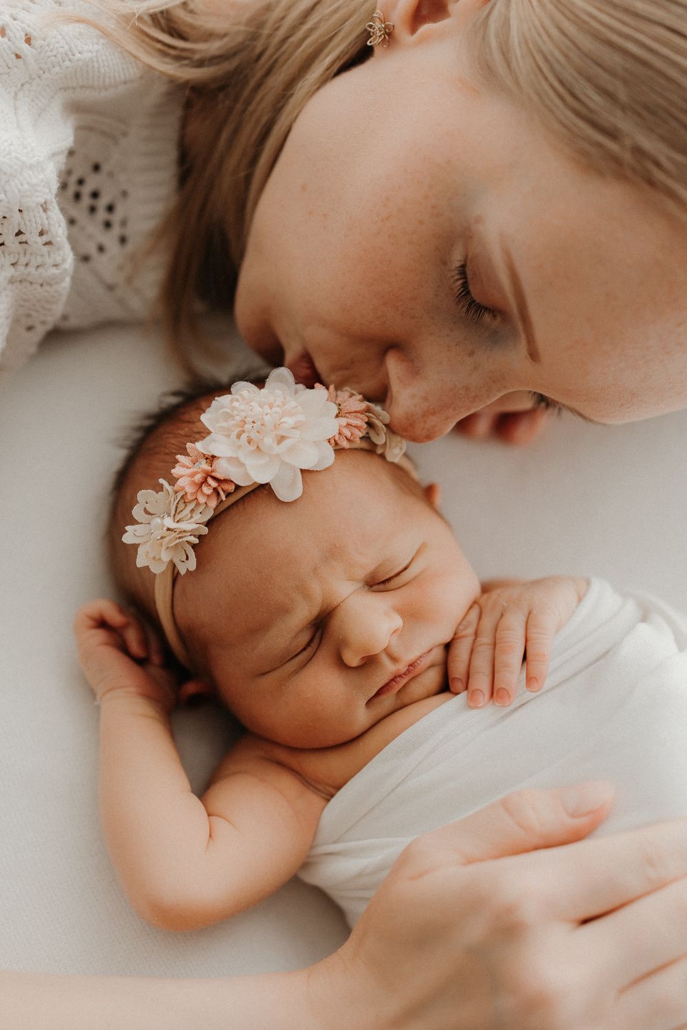 A mother tenderly kisses her sleeping newborn baby girl wearing a floral headband, wrapped in white.
