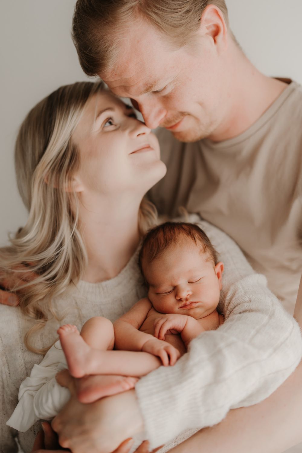 A couple tenderly touches foreheads while cradling their sleeping newborn baby wrapped in a white blanket.