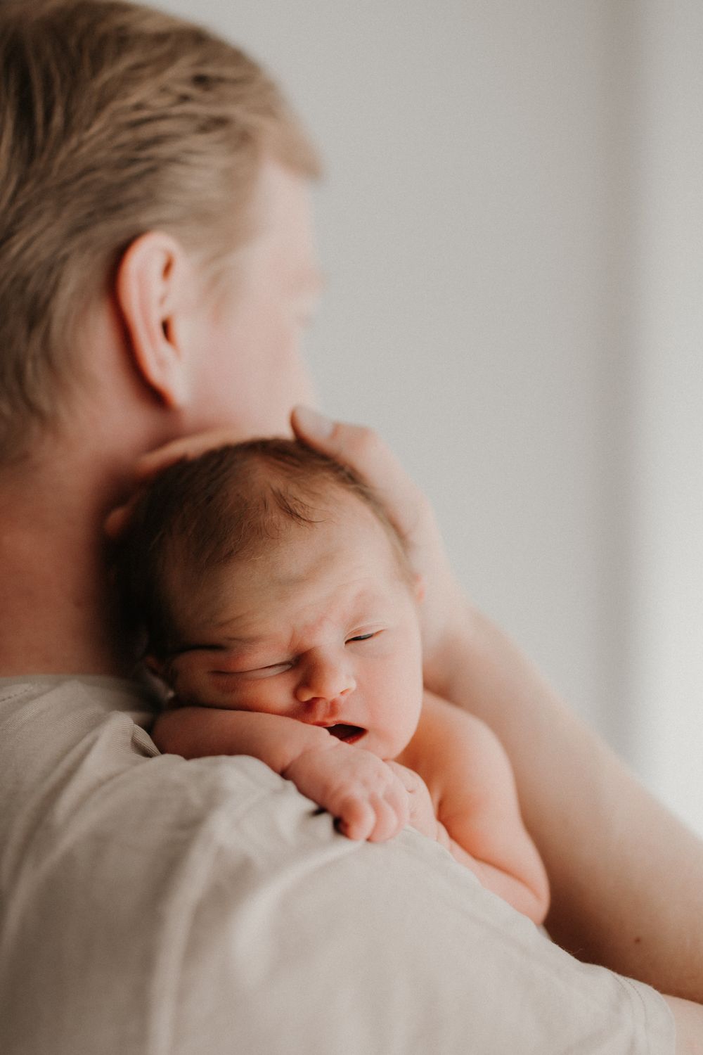 A parent tenderly holds a sleeping newborn baby against their shoulder in a soft, warmly lit intimate moment.