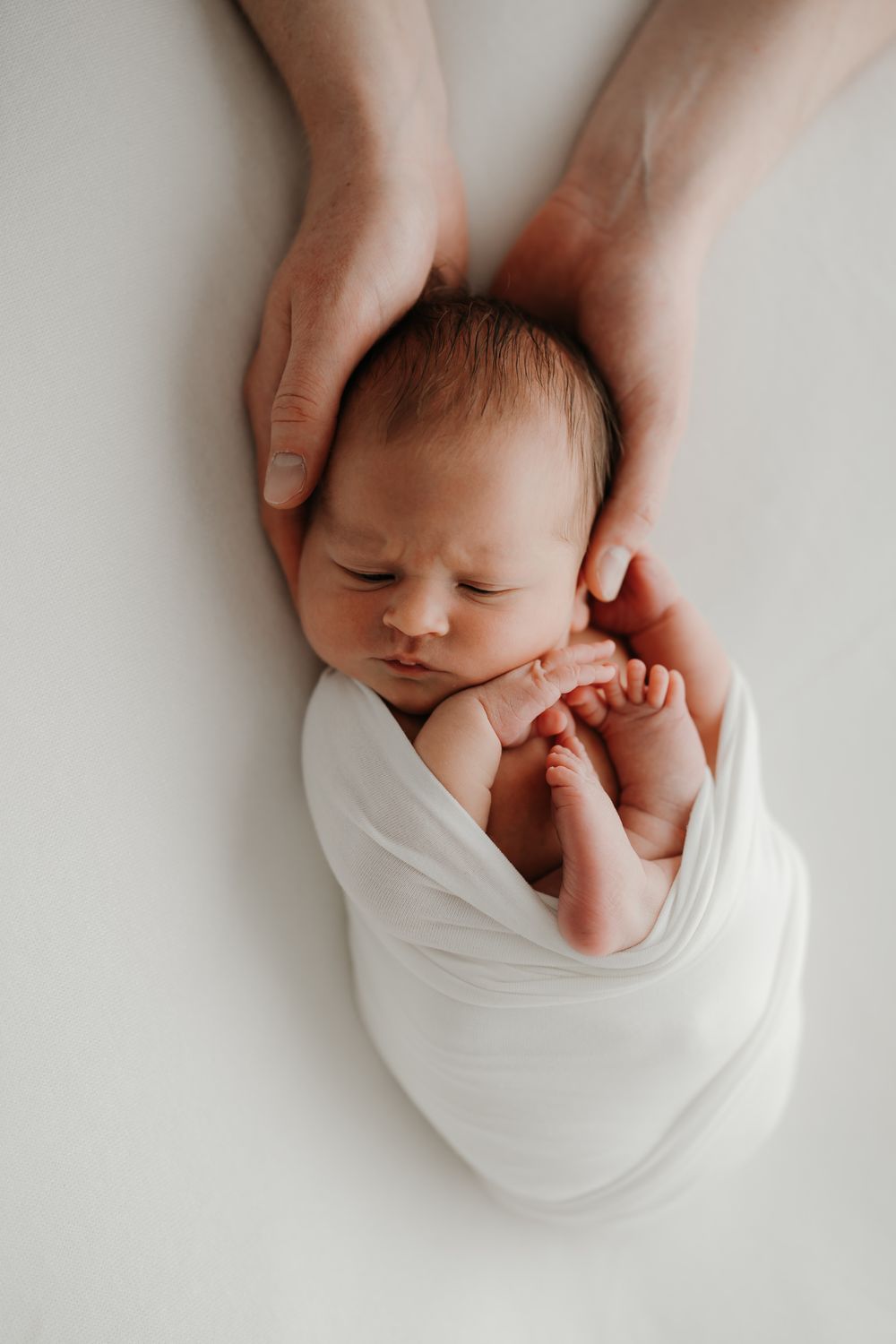 Newborn baby swaddled in white wrap, sleeping peacefully while adult hands gently cradle the infant's head.