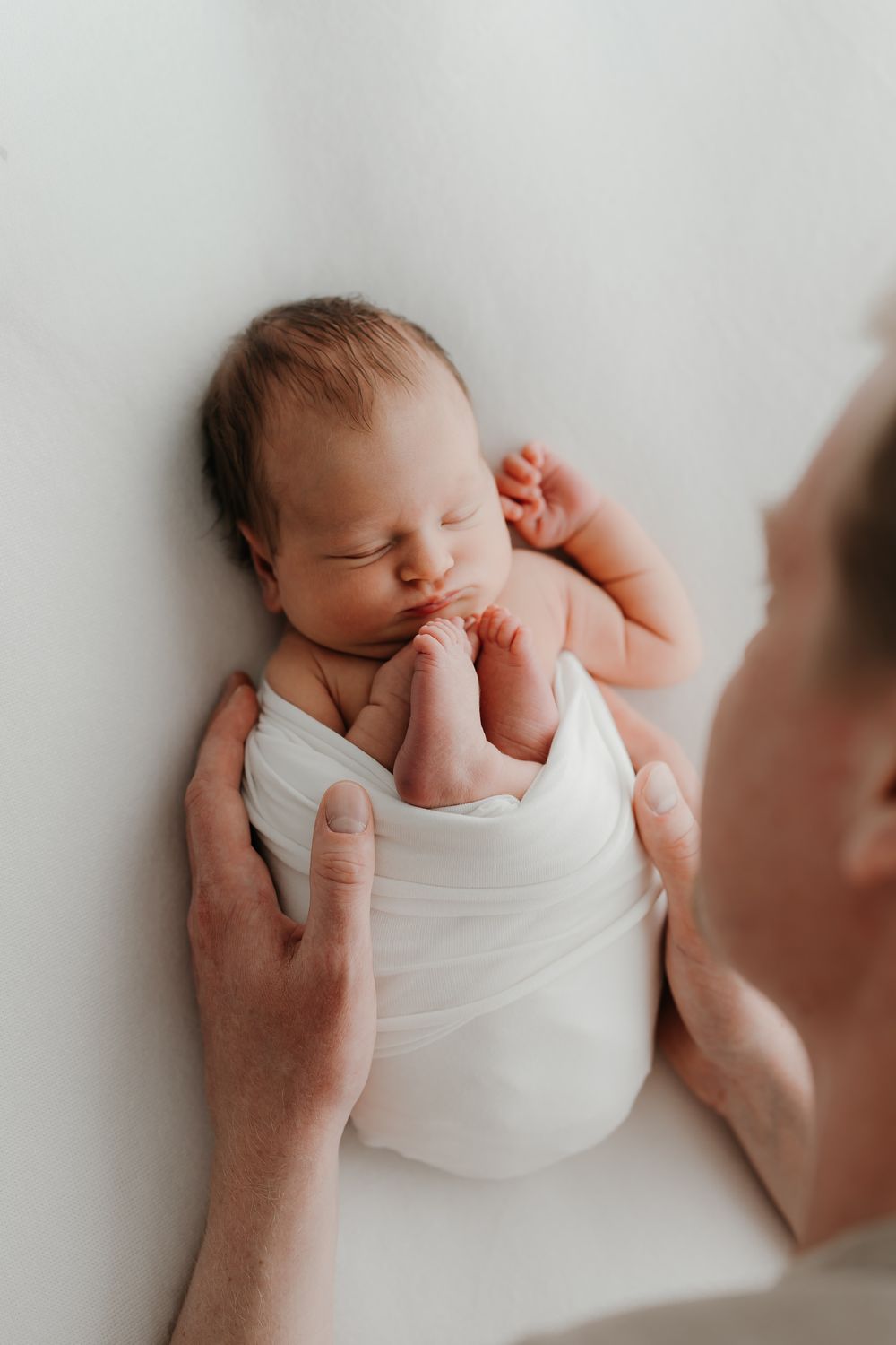 A newborn baby swaddled in white sleeps peacefully while a parent gently holds them from above.
