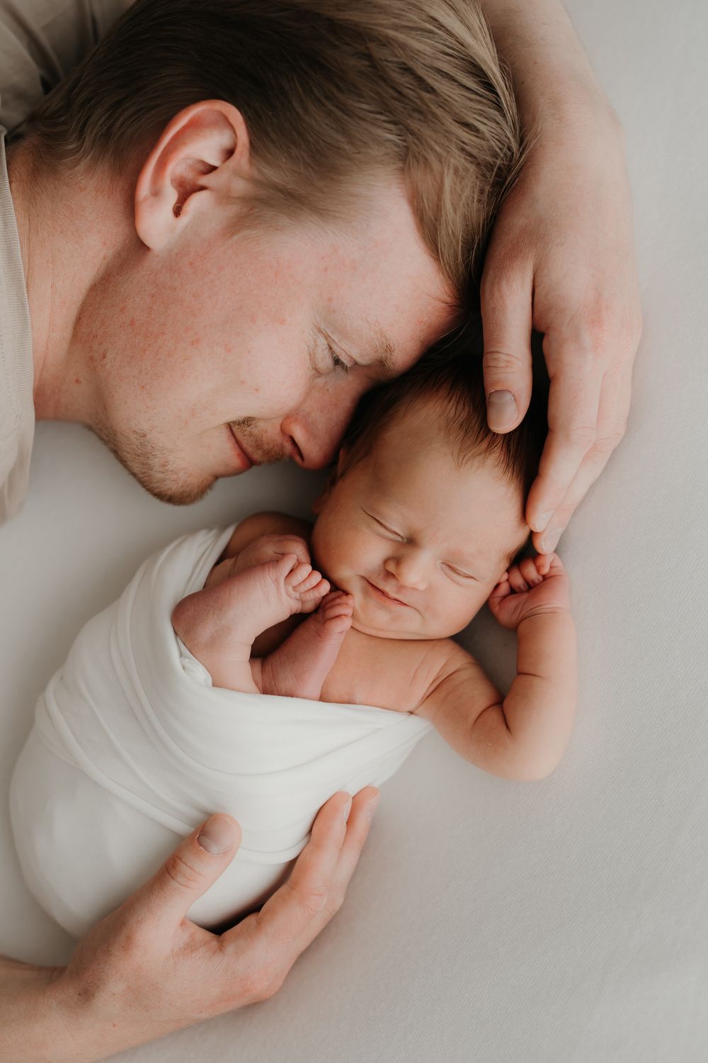 Father tenderly resting beside sleeping newborn baby wrapped in white, sharing a peaceful bonding moment.