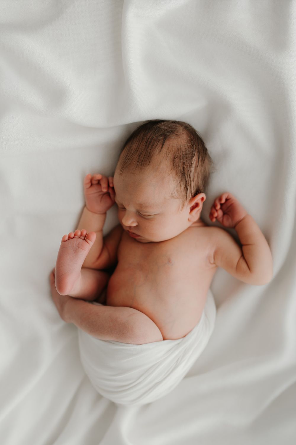 Peaceful newborn baby sleeping on white fabric, wrapped in a white swaddle with tiny hands raised.