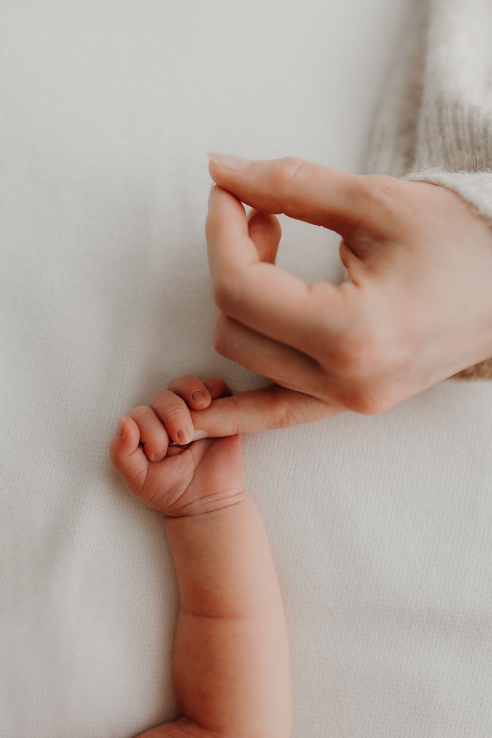 A mother's hand gently holds her newborn baby's tiny fingers on a soft white blanket.