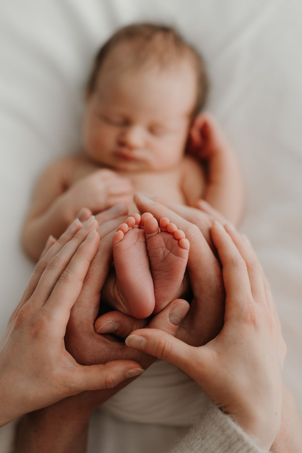 Parents gently cradling their sleeping newborn baby's tiny feet in their hands on a white blanket.