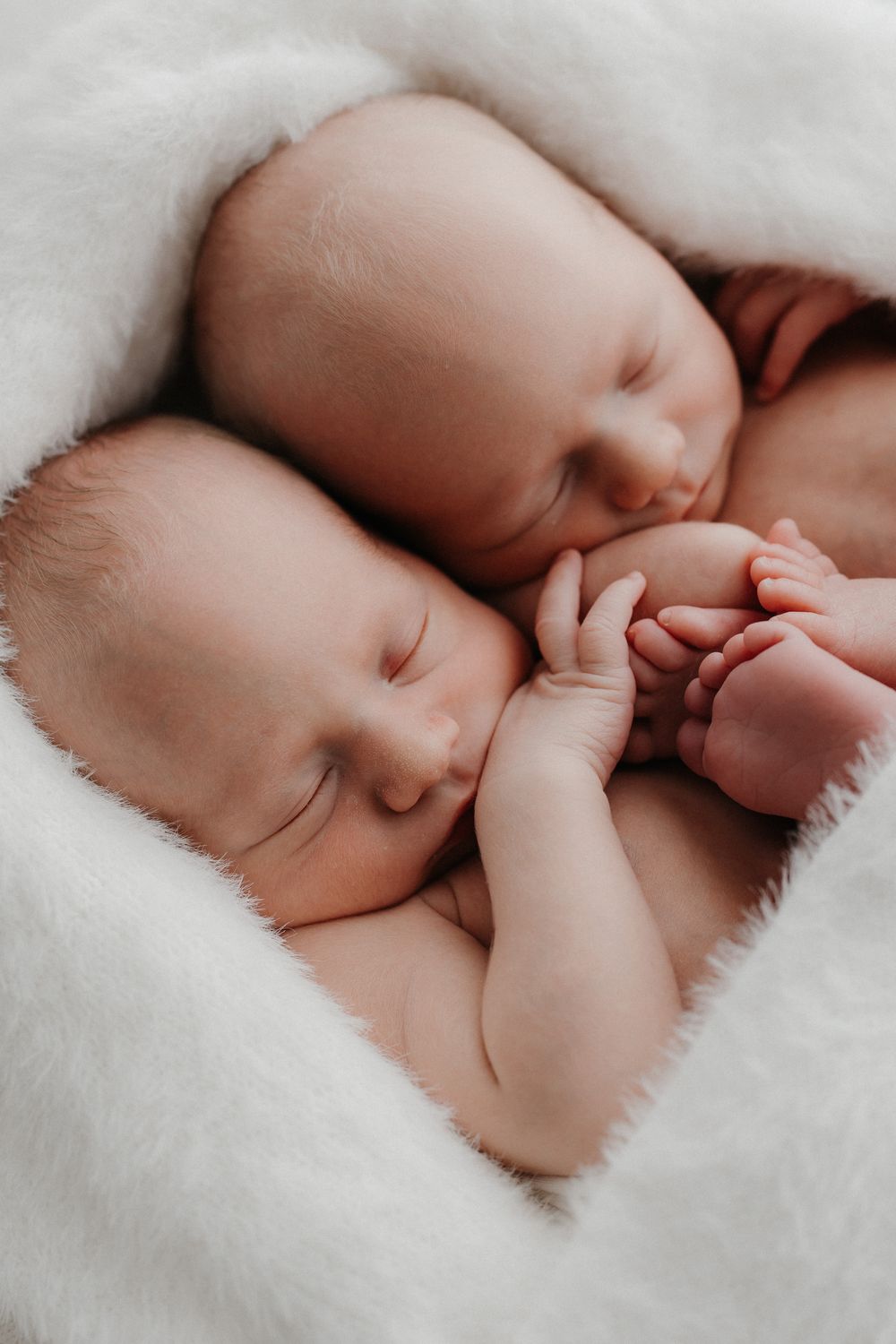 Two sleeping newborn twins cuddle together on a soft white fluffy blanket, holding hands in a peaceful moment. Newborn-kuvaus kaksoset.