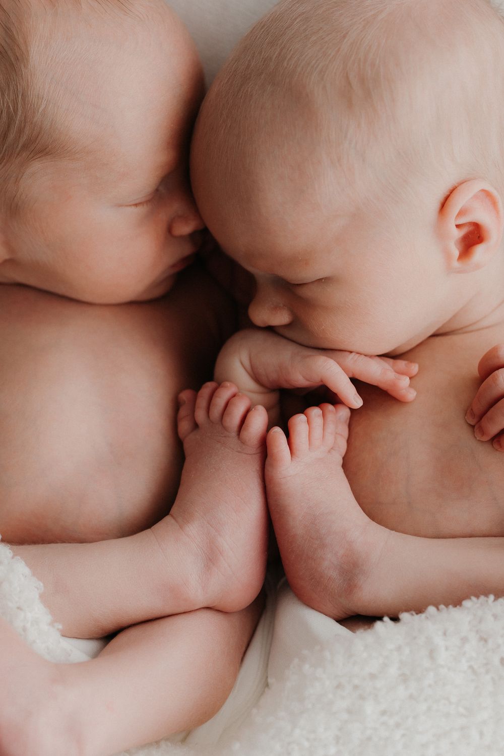 Two newborn twins sleeping closely together, holding each other's tiny feet on a soft white blanket. Newborn-kuvaus kaksoset.