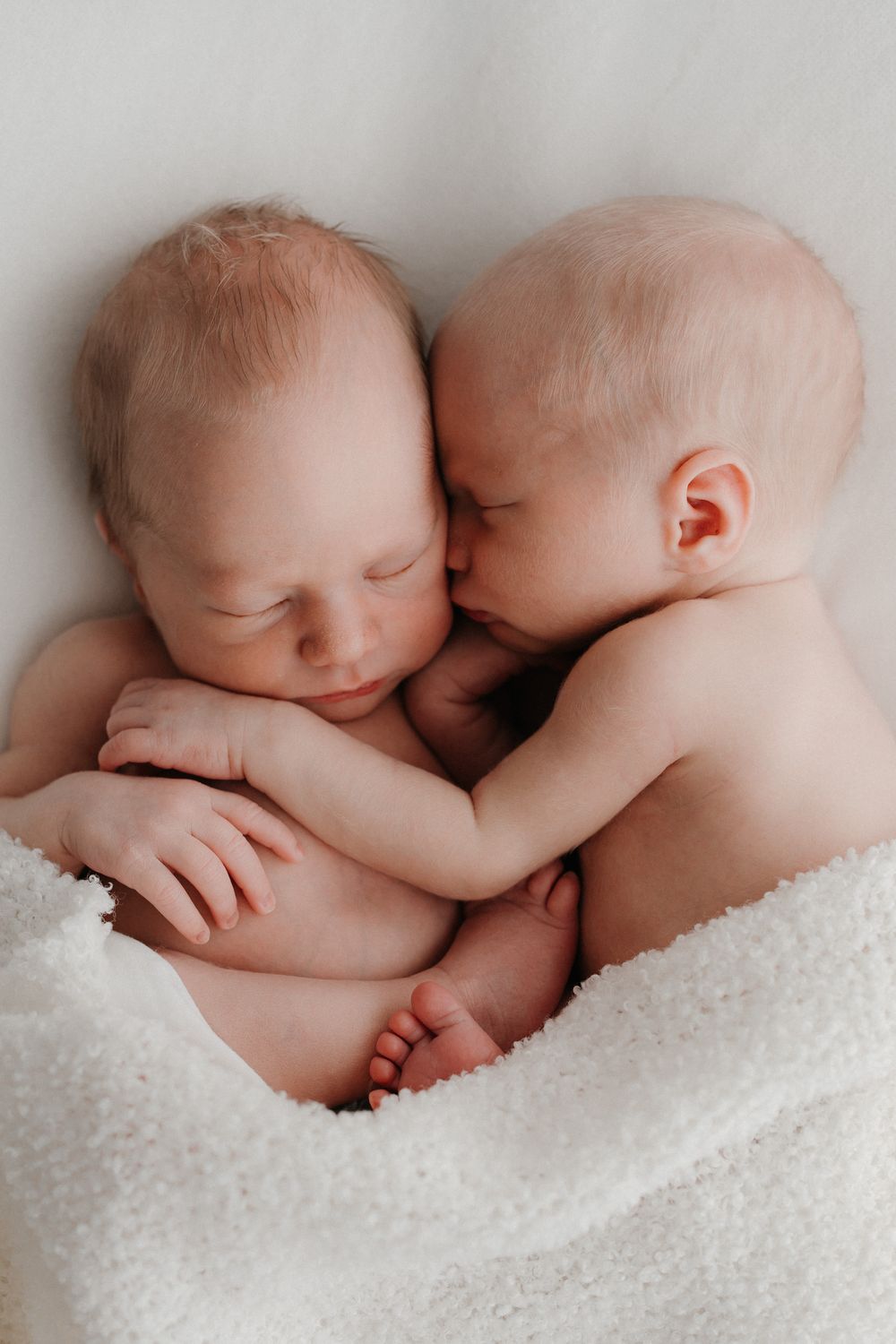 Two newborn twins cuddling together on a white blanket, one gently kissing the other in a tender moment. Newborn-kuvaus kaksoset.