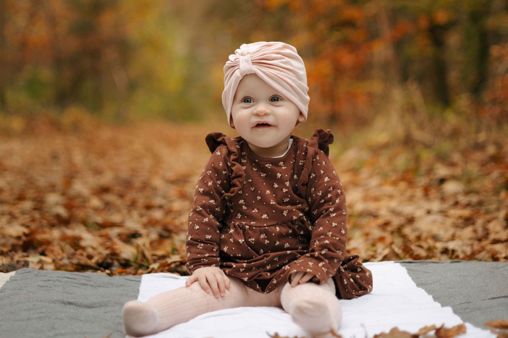 Baby in brown polka dot dress and pink headwrap sitting on blanket amid fall foliage.