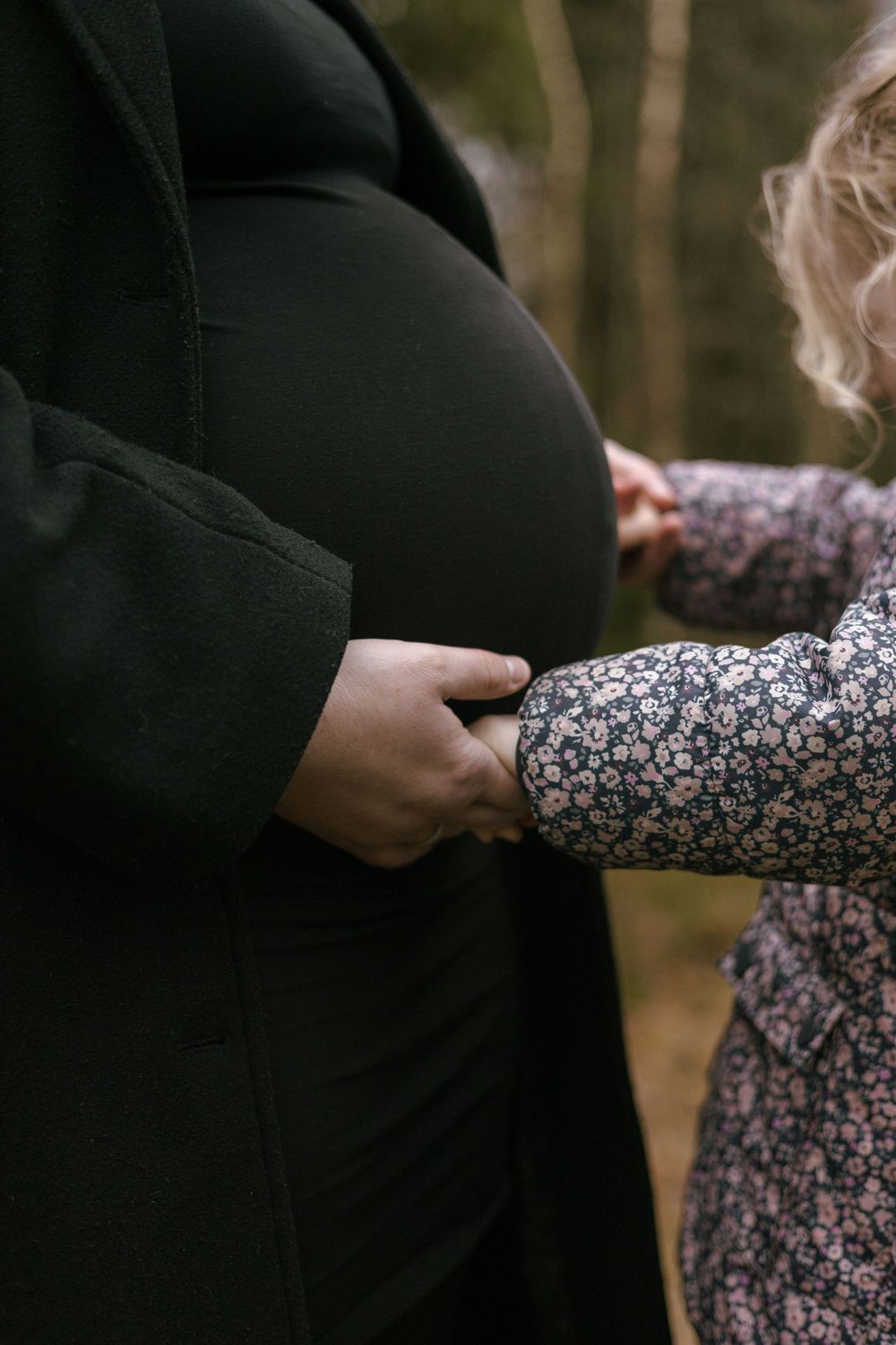 Someone in a floral dress reaches out to touch a pregnant person wearing black in an intimate outdoor moment.