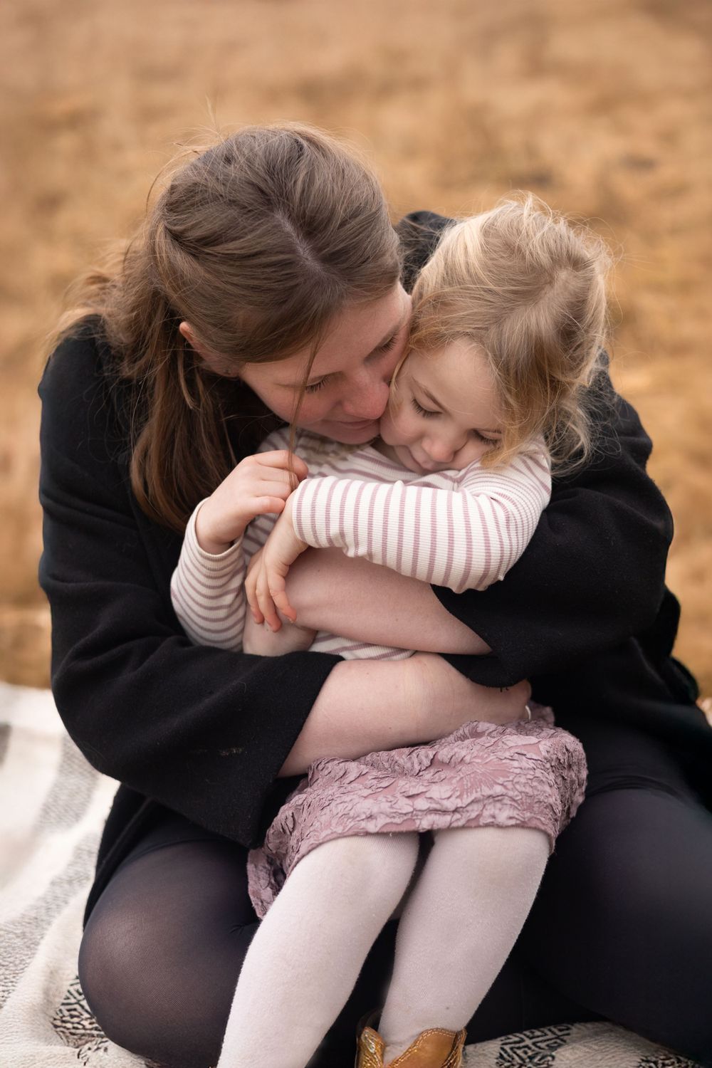 A tender moment of comfort and affection between parent and child sitting together on a blanket outdoors.