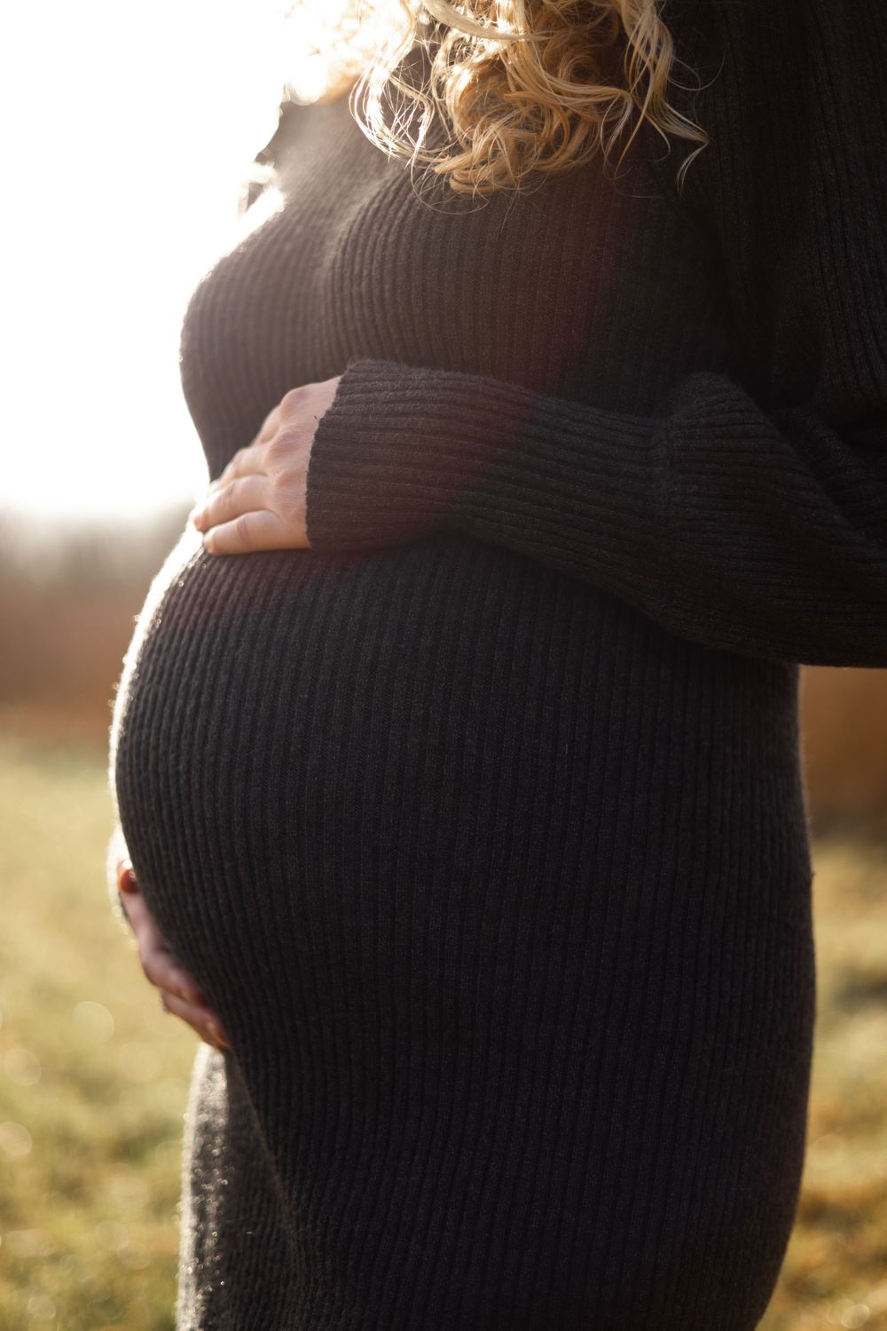 A silhouetted figure in a dark ribbed sweater cradles their pregnant belly in warm sunlight.
