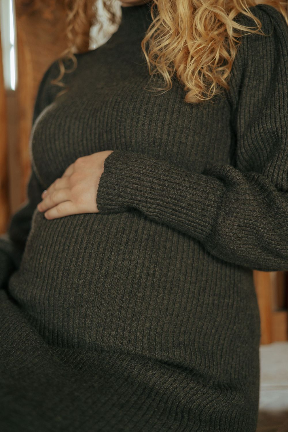 Close-up of pregnant woman wearing cozy black ribbed sweater dress showing baby bump in warm lighting.