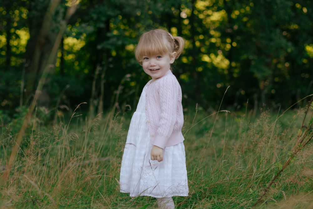 Child wearing delicate pink and white dress poses in a sunlit meadow.