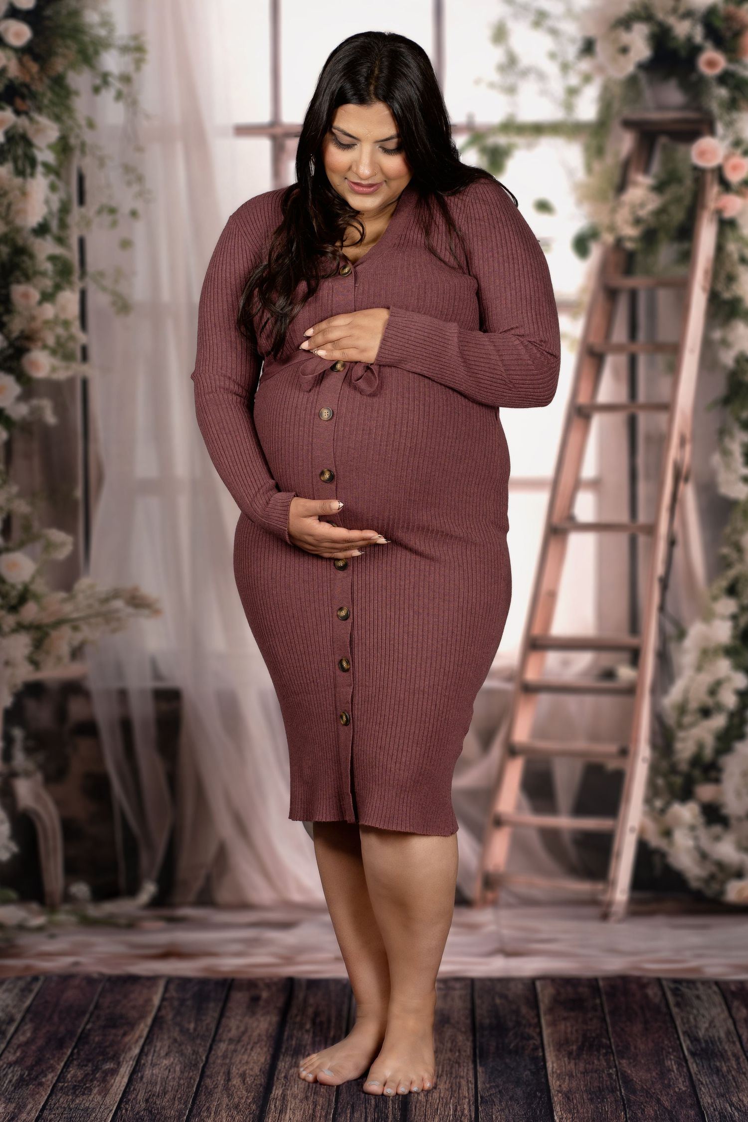 Pregnant woman in maroon ribbed sweater dress poses barefoot against floral backdrop.