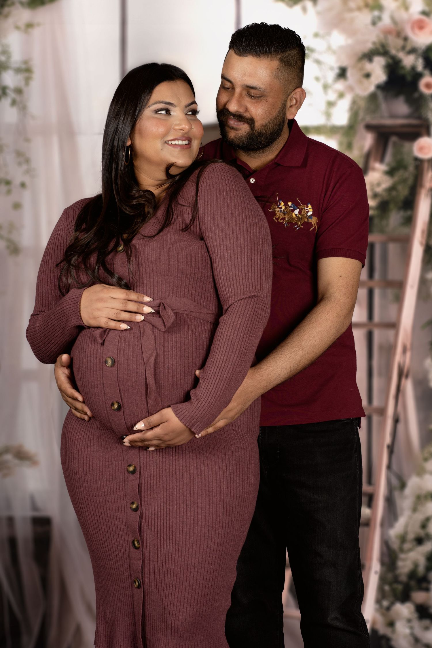 Maternity photo featuring couple in matching burgundy outfits among floral decorations.