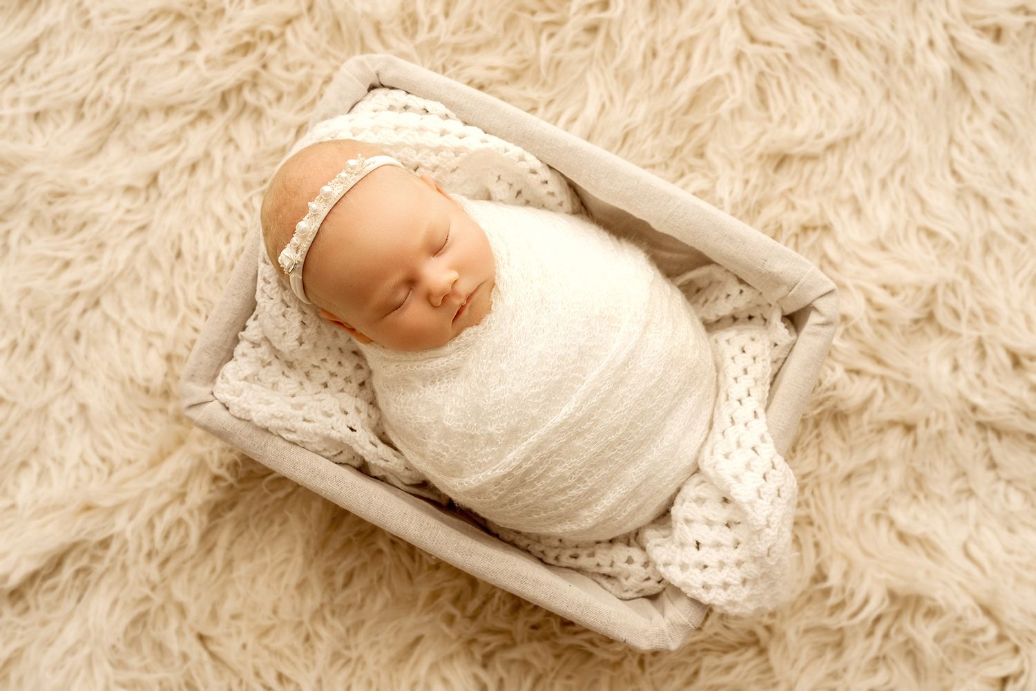 Newborn baby wrapped in a lace white wrap lying in a rustic basket on cream fluffy rug