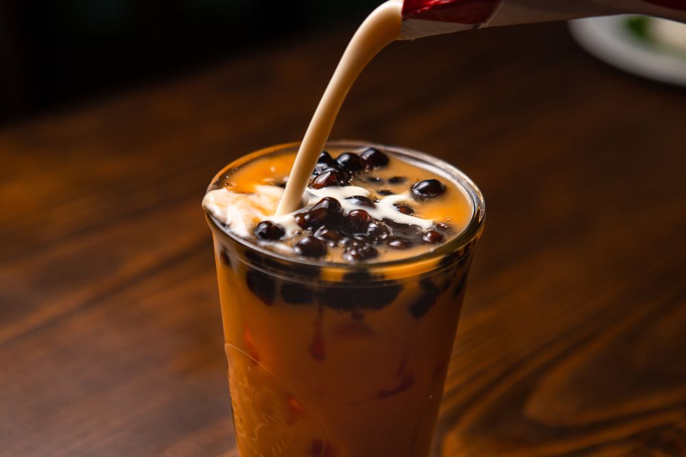 Rich milk cream pouring into cold tea with dark boba pearls on a wooden table.