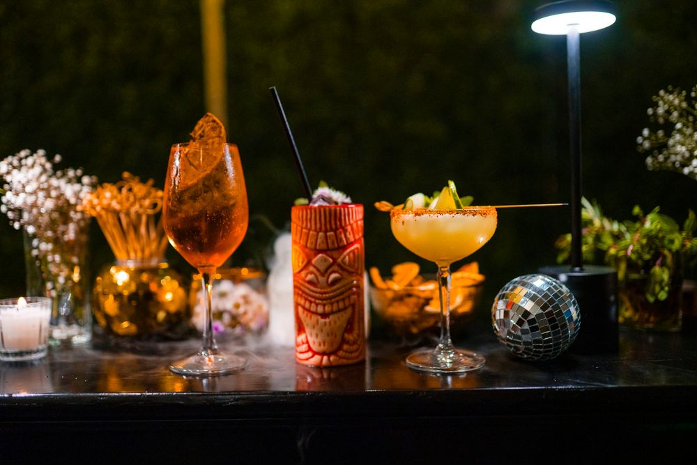 Creative cocktails with orange garnishes arranged on a dark bar under moody lighting with glowing tiki glassware.