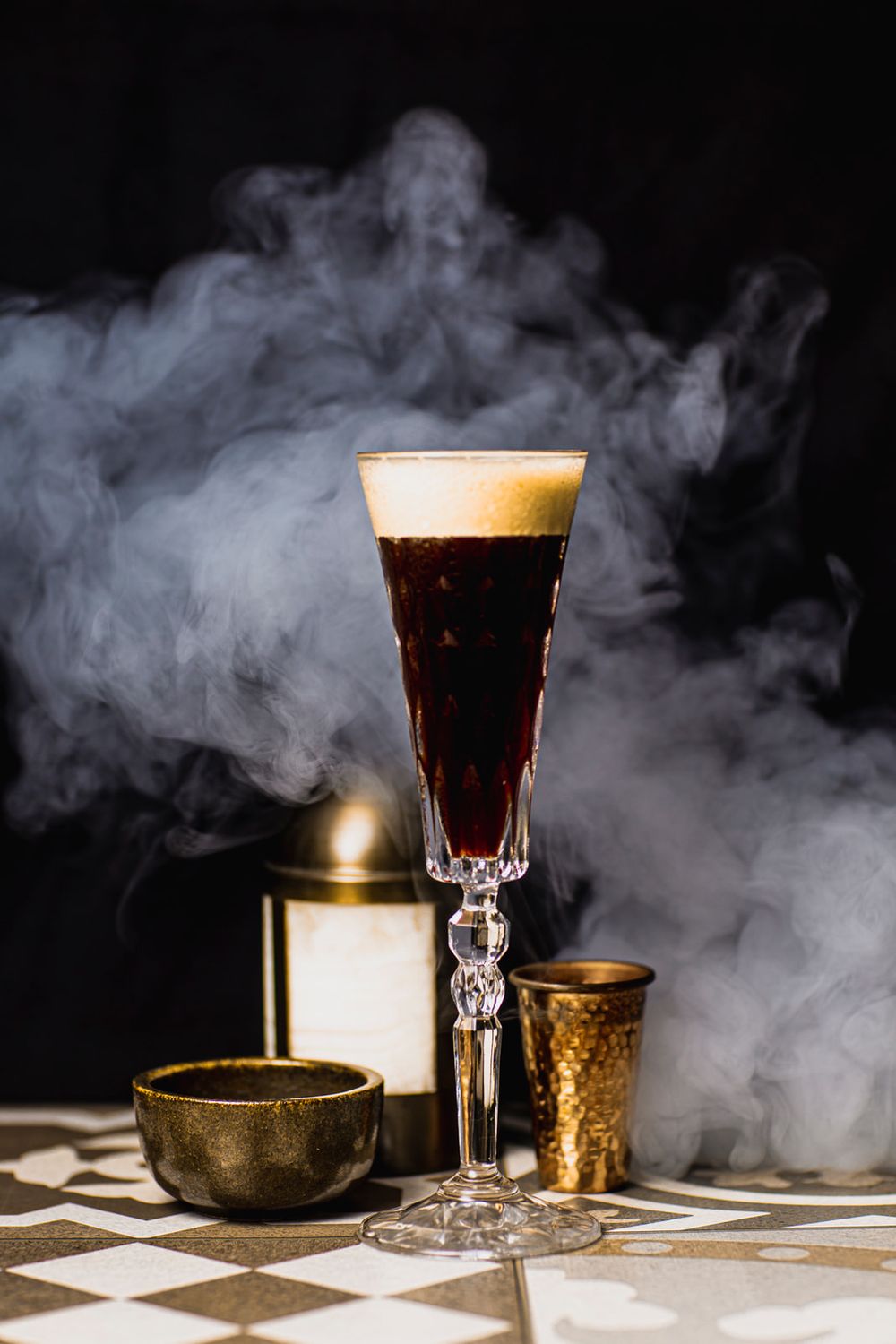 A dark beer with frothy head in an elegant crystal glass sits on checkered surface amid moody smoke and golden accessories.