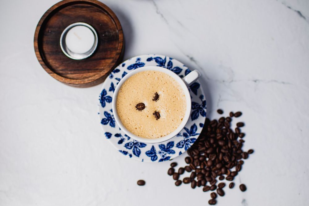 Coffee cup with frothy latte on blue and white china with scattered coffee beans on marble surface.