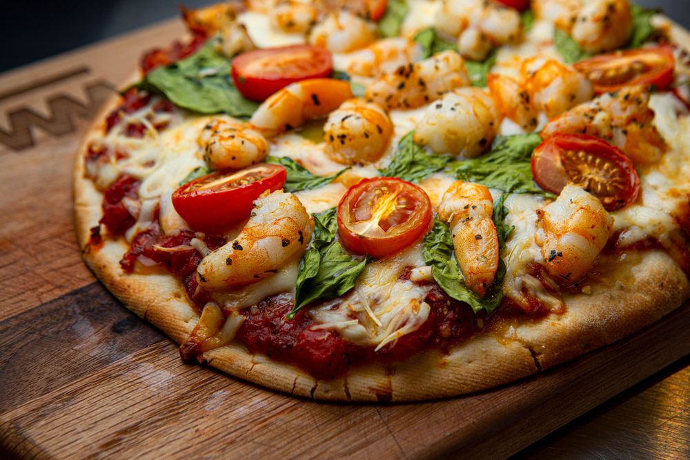 Gourmet pizza topped with grilled shrimp, cherry tomatoes, spinach leaves, and melted cheese on a wooden serving board.