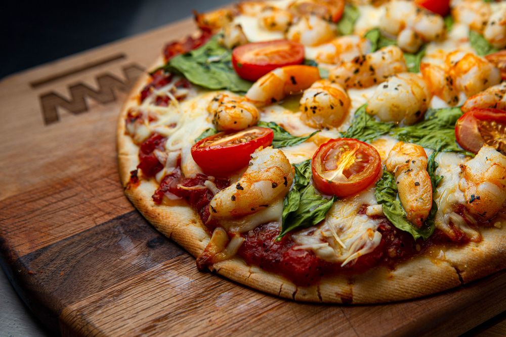 Fresh gourmet pizza topped with shrimp, cherry tomatoes and arugula on wooden cutting board.