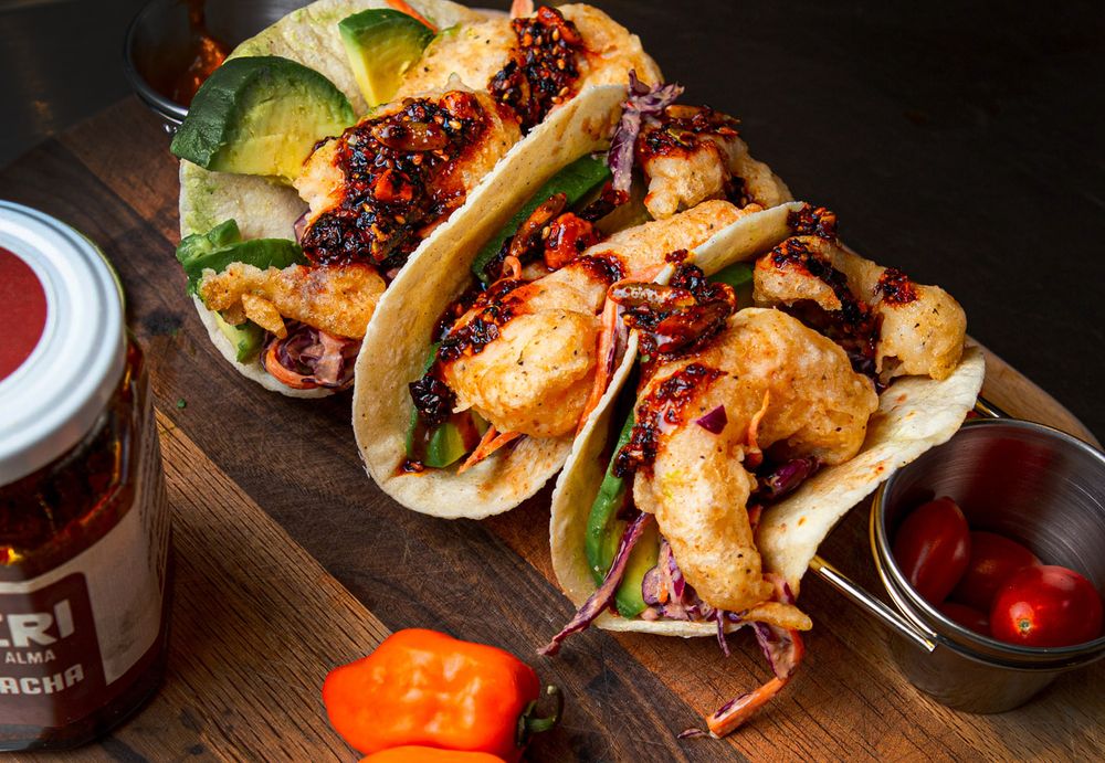 Three grilled fish tacos topped with fresh avocado, cabbage slaw and spicy sauce served on a wooden board with condiments.