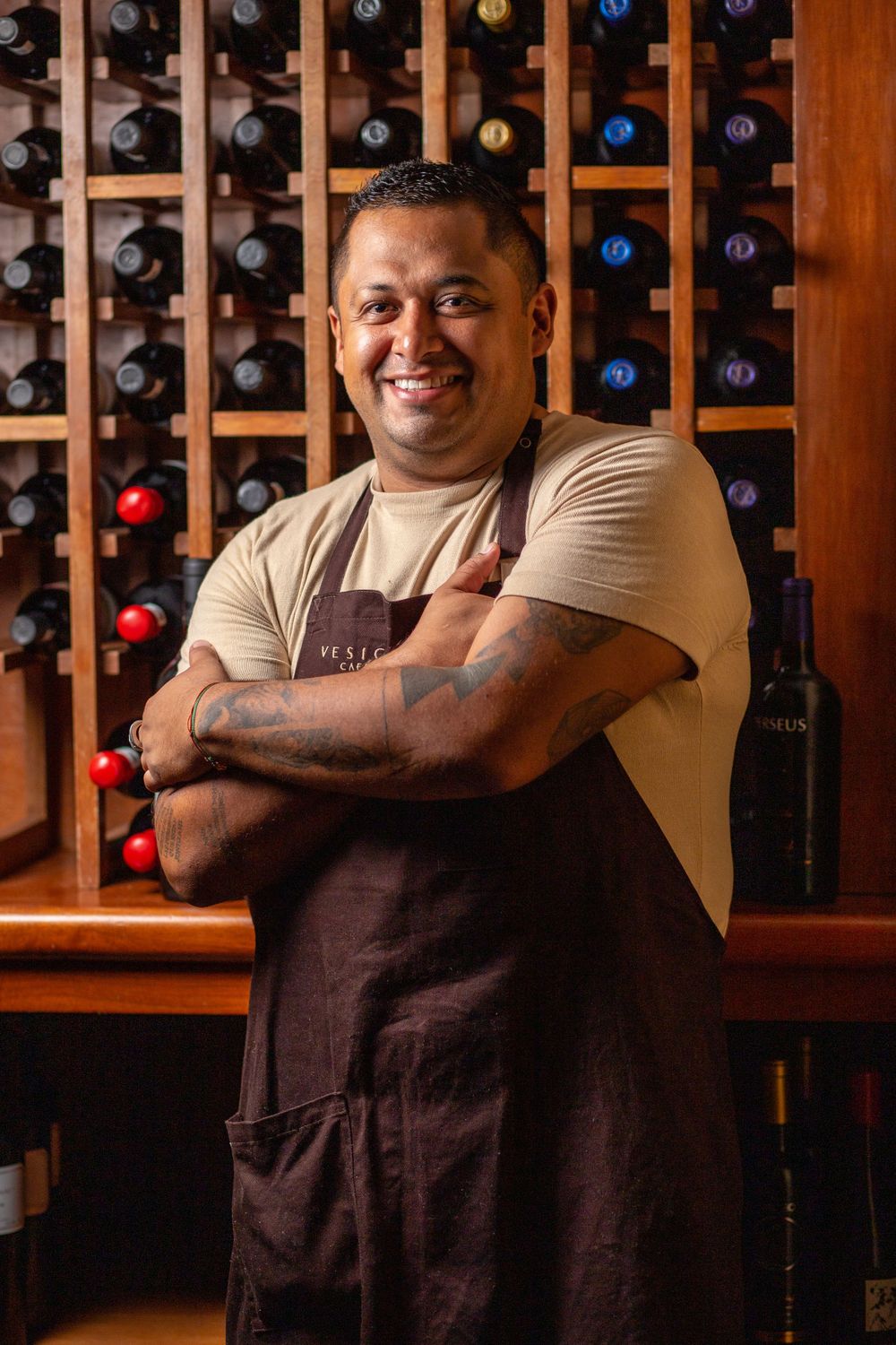 Restaurant staff member wearing brown apron stands smiling with crossed arms in front of wine rack display.