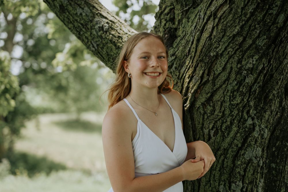 A woman in white dress poses cheerfully by a large tree trunk.