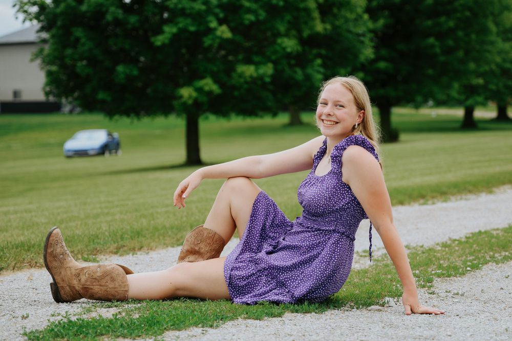 Casual outdoor portrait sitting on gravel path wearing boots and purple dress.