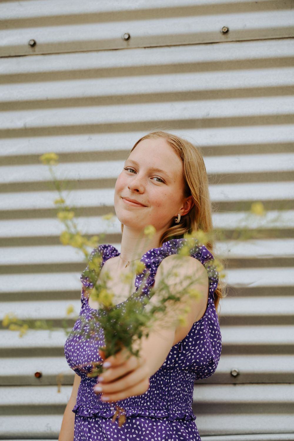 Holding wildflowers while wearing purple dress against metal siding background.