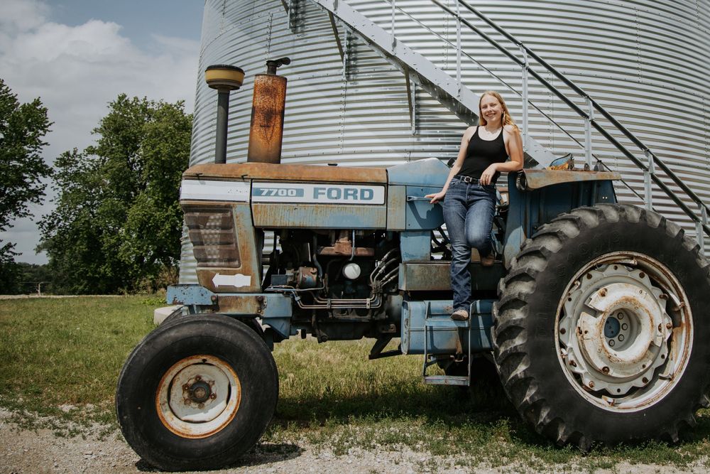 A vintage Ford tractor sits next to a large metal grain silo on a sunny day in the countryside.