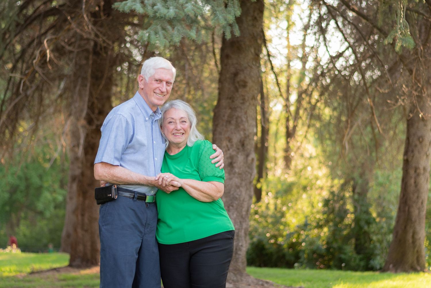 Two people in casual attire share a tender moment while sitting outdoors in a sunlit wooded area.