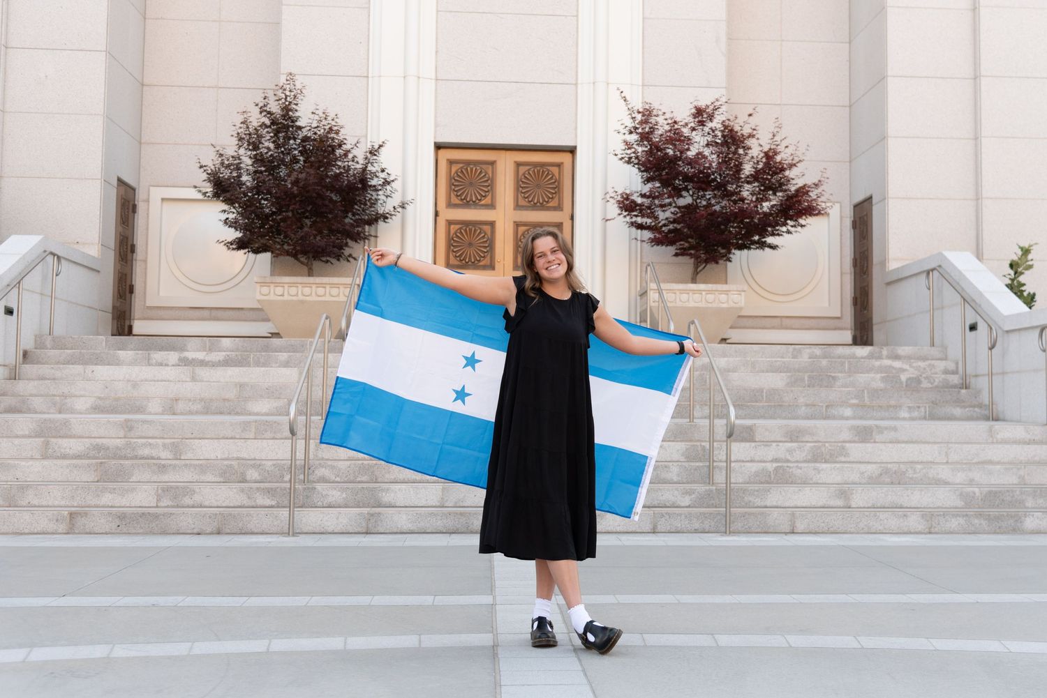 Person in black dress holding blue shopping bags stands on marble steps outside grand stone building entrance.