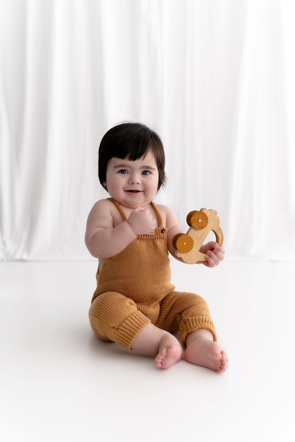 A baby in mustard yellow romper sitting with teddy bear against white background.