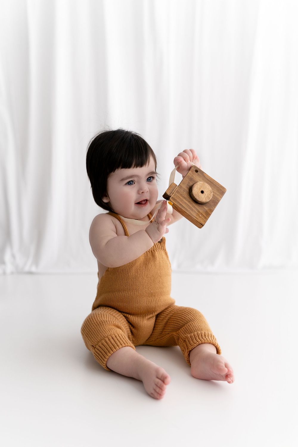 Baby in mustard romper plays with wooden toy against white backdrop.