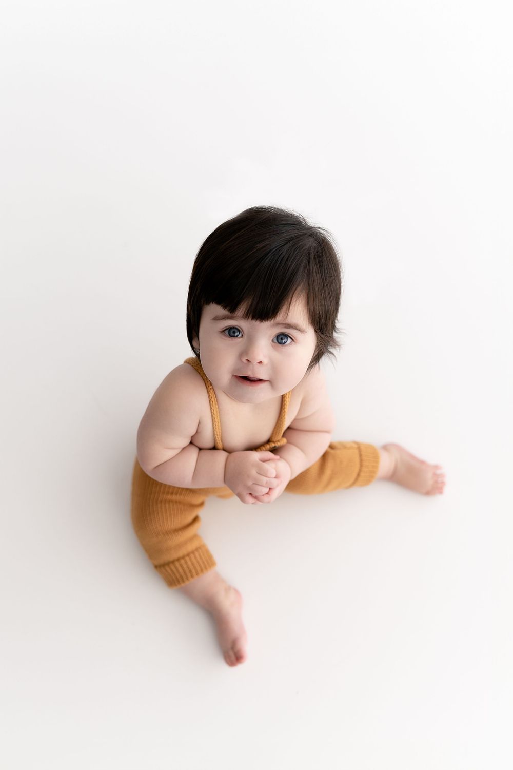 Baby in orange knitwear sits on white floor during minimalist photo session.