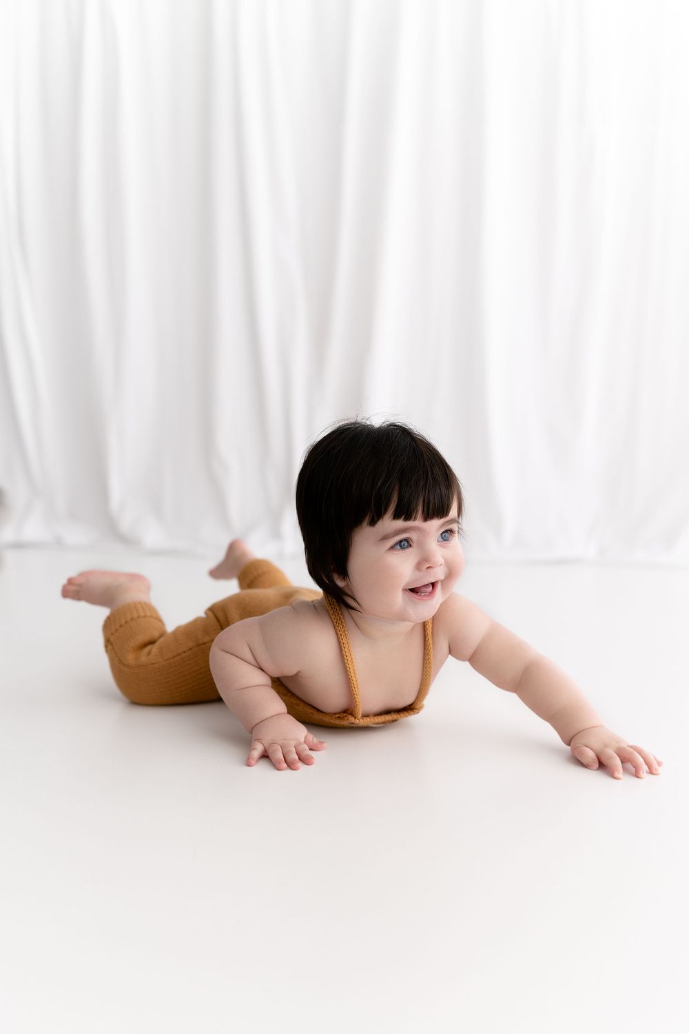 Baby wearing mustard yellow romper lying on stomach on white floor against white curtain background.