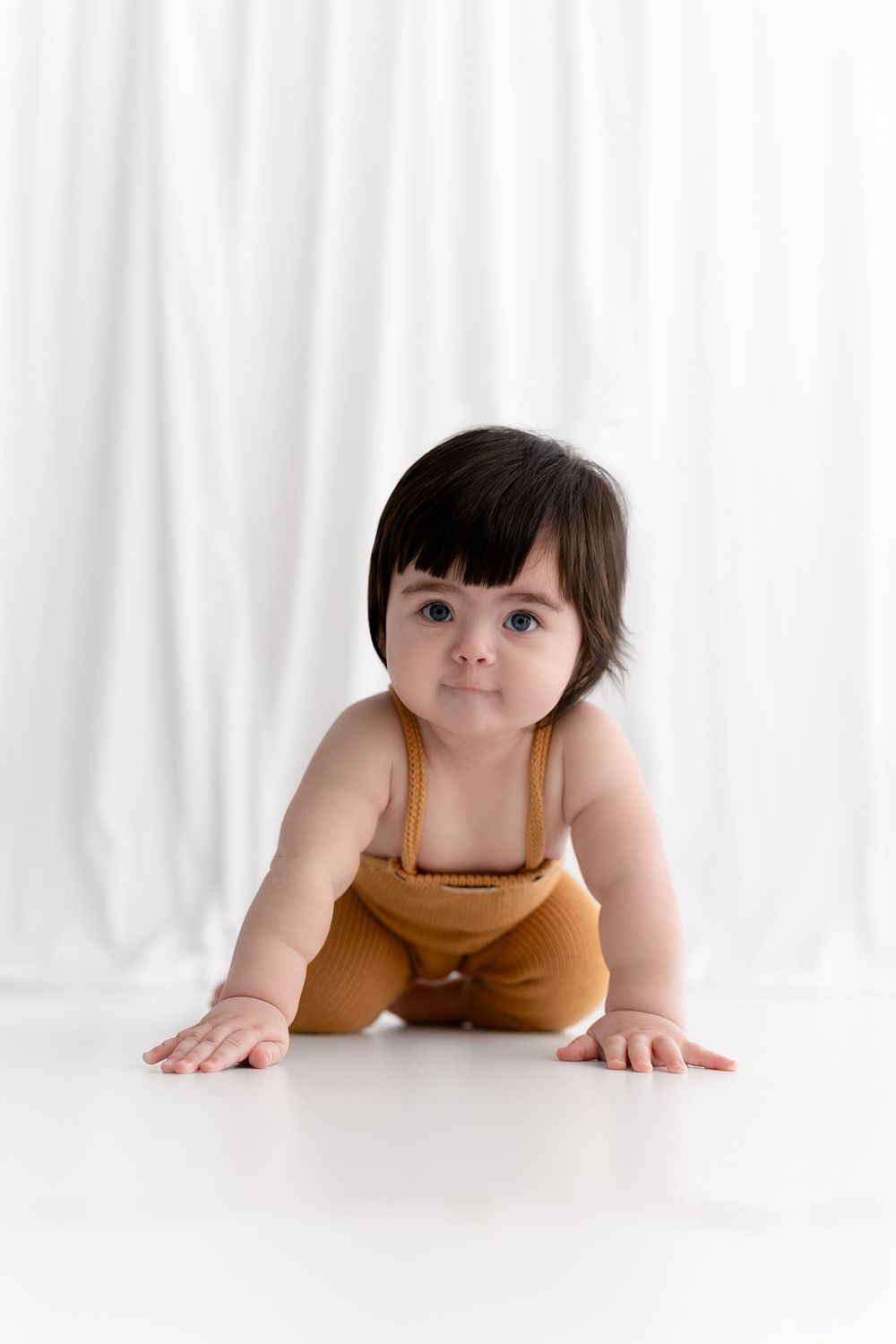 Smiling baby in orange romper crawls on white floor during studio photoshoot.