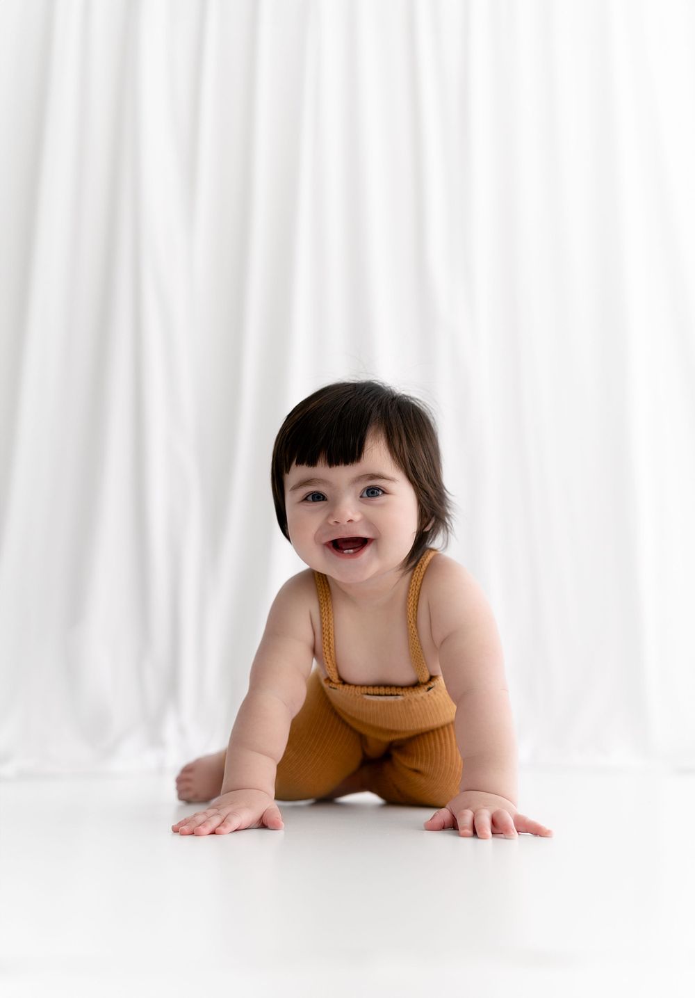 Happy baby in mustard romper crawling on white floor with big smile.