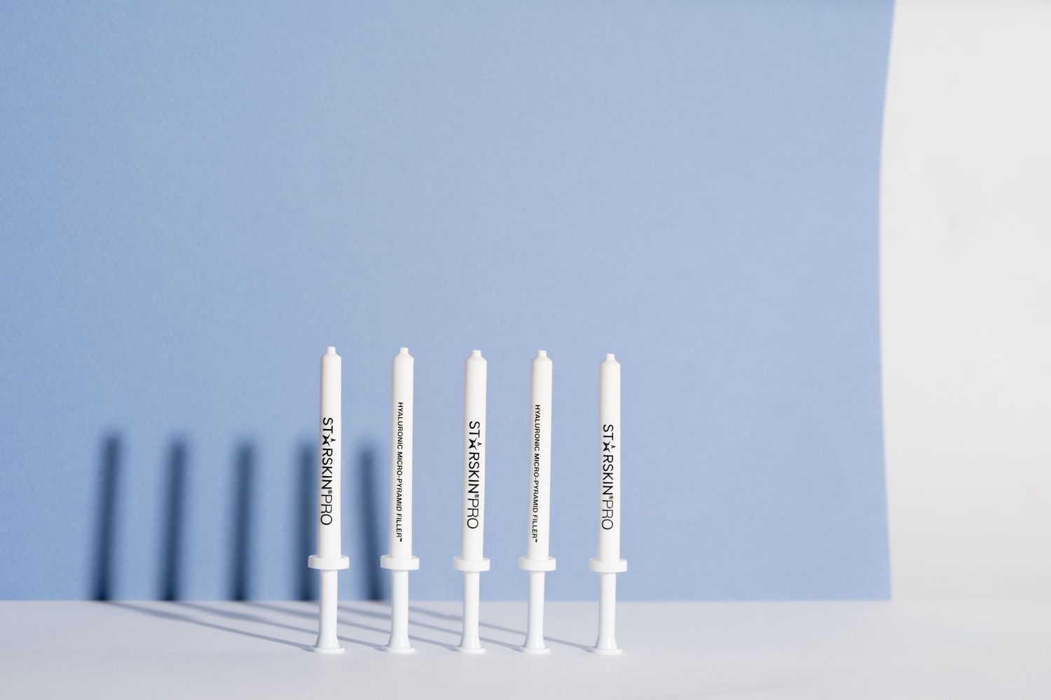 Een lichte productfoto van een beautyproduct, het product staat een paar keer tegen een blauwe achtergrond met een harde schaduw.