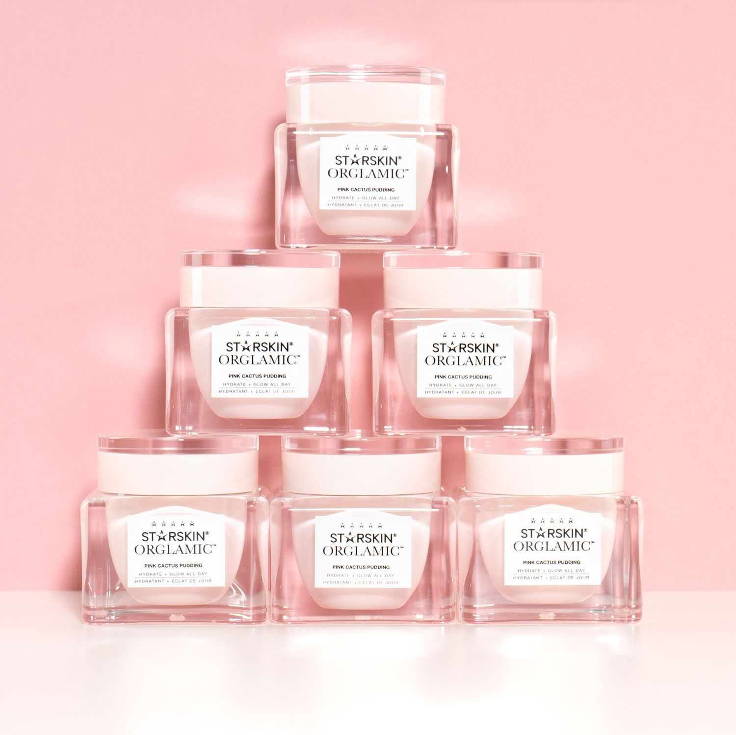Een lichte productfoto van een beautyproduct, de potjes zijn als piramide gestapeld tegen een roze achtergrond.