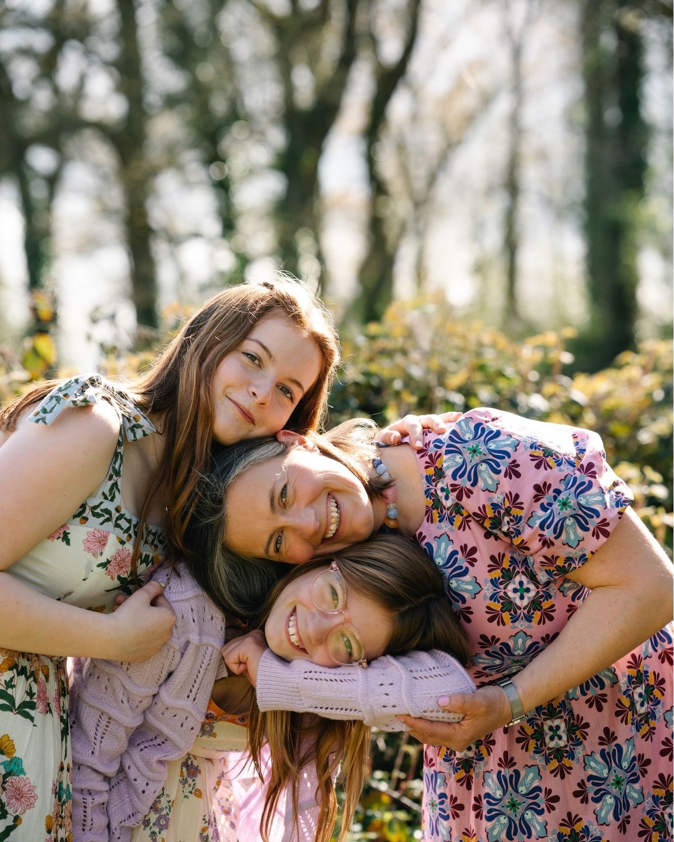 Three smiling young women in floral dresses huddle together outdoors, posing playfully among spring trees.