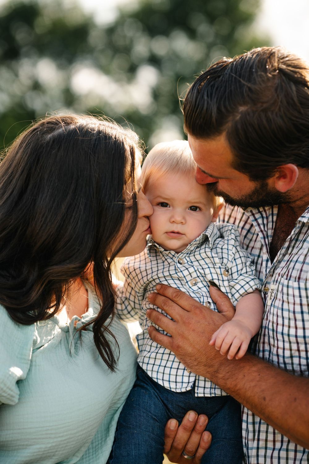 Family of three showing tender moments of affection with parents kissing young child wearing plaid shirt outdoors.
