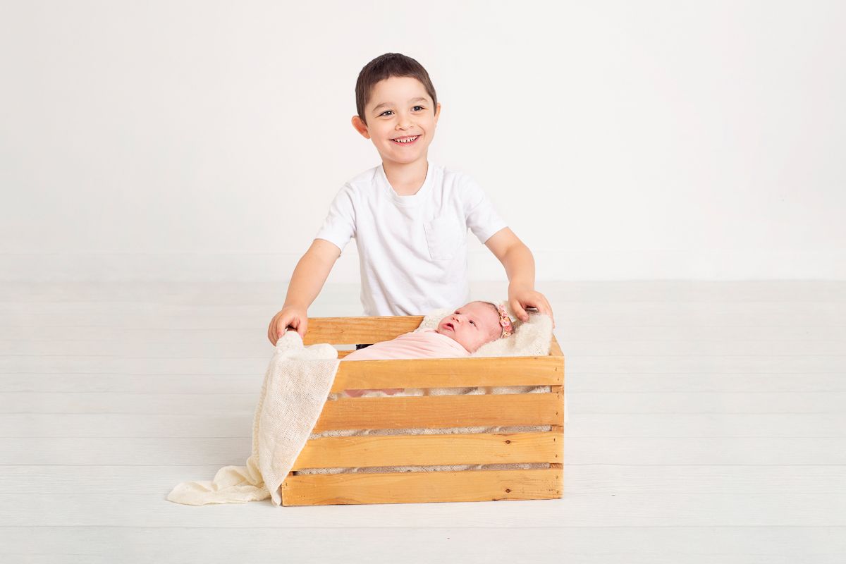 smiling little brother, school age boy sits beside wooden prop where newborn baby sister in laying in