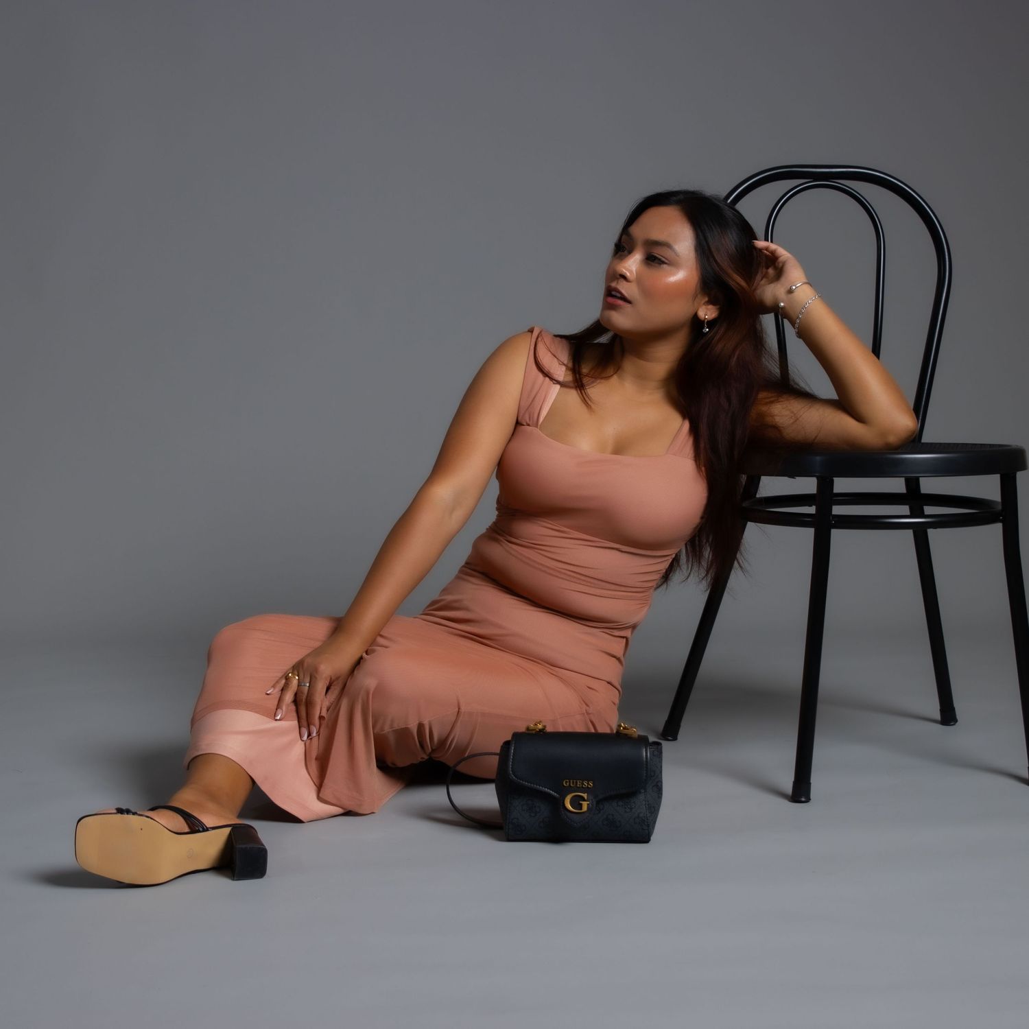 Fashion portrait in dusty rose dress with black handbag and chair against grey backdrop.