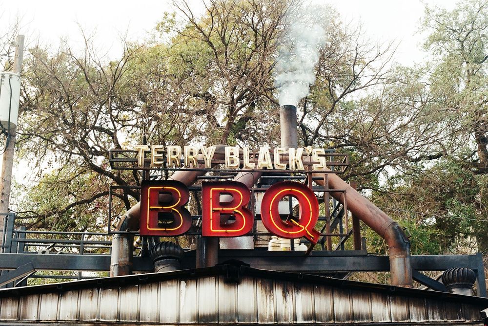 Neon sign for Terry Black's BBQ restaurant with smoke rising against tree-lined sky in Austin, Texas.