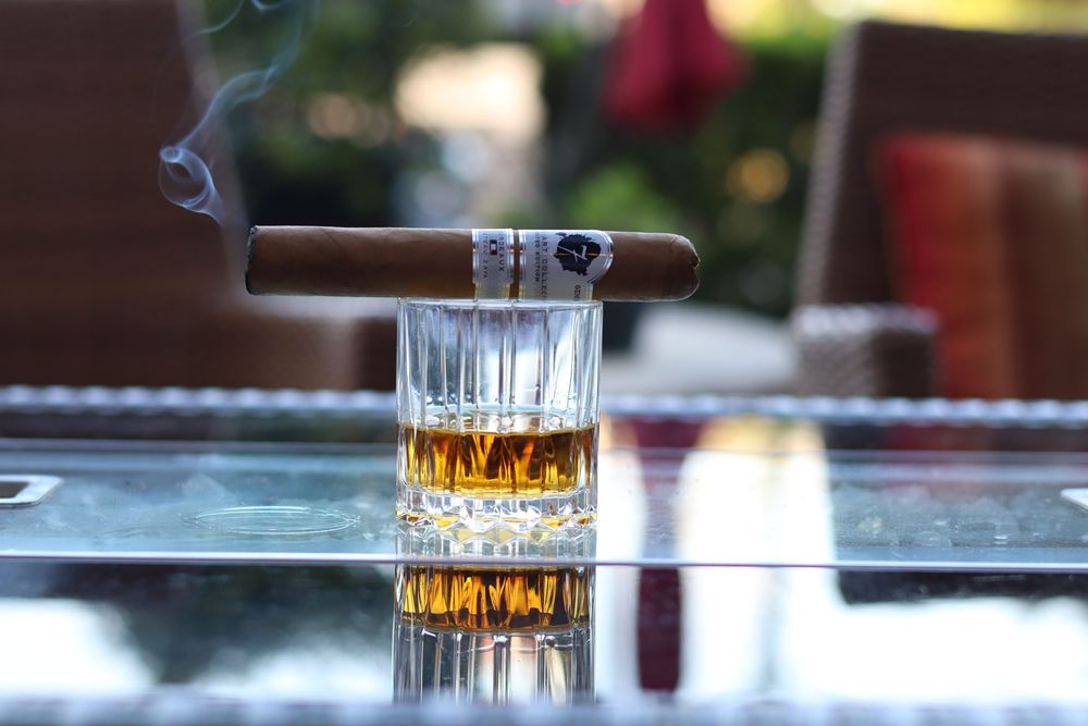A lit cigar resting on a crystal whiskey glass filled with amber spirits on a glass table outdoors.